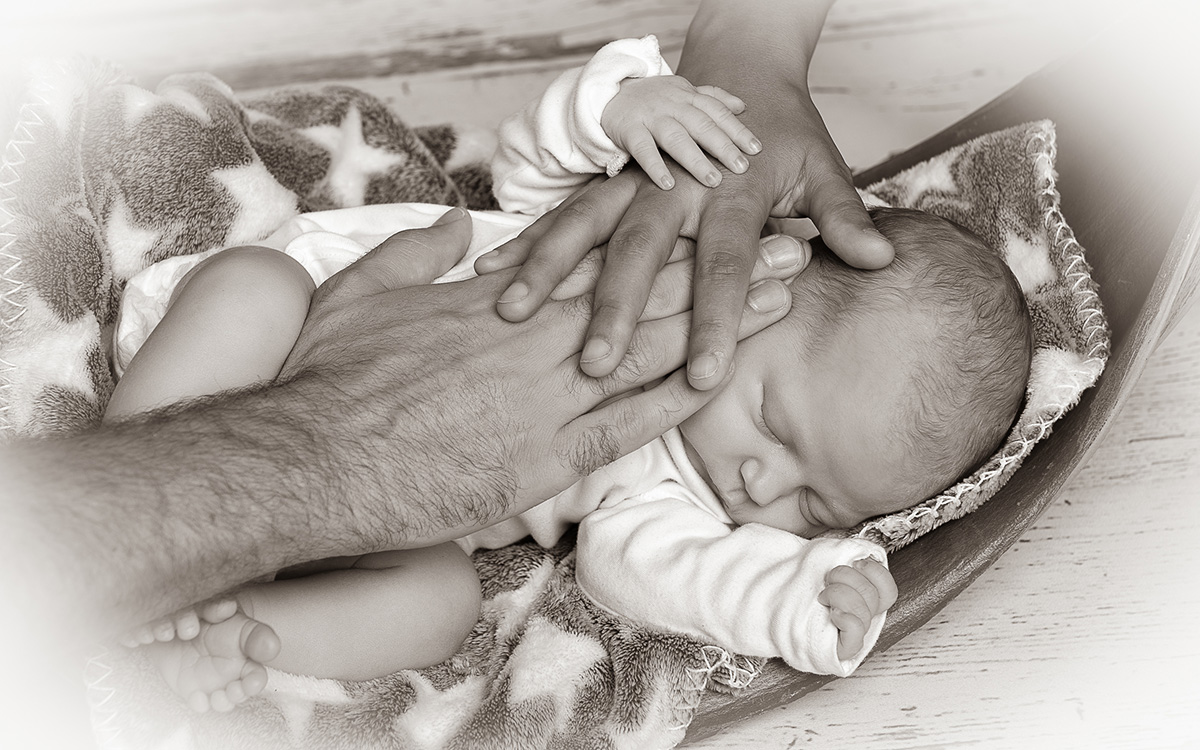 Schwarz-weiß Bild eines Babys, auf das die Hände der Eltern gelegt sind. Das Baby greift nach der Hand der Mama.